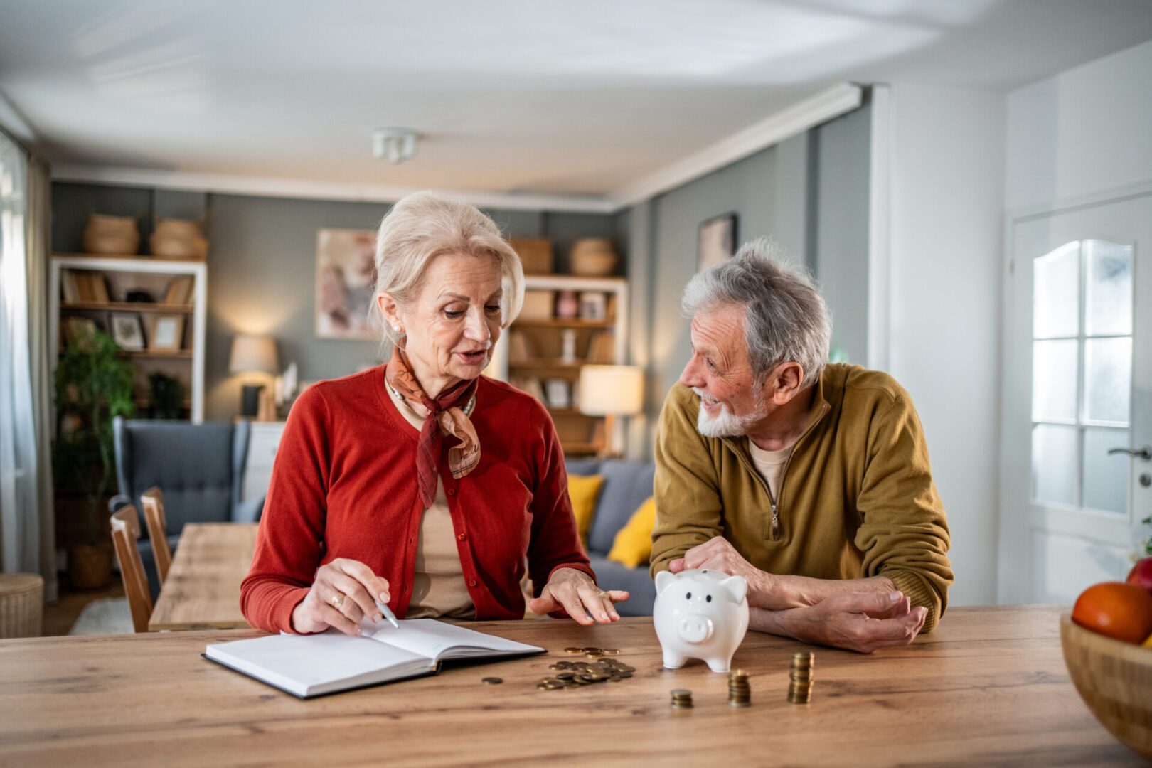 Happy senior couple calculating their expenses and managing their savings at home, using a piggy bank and a notebook to keep track of their finances